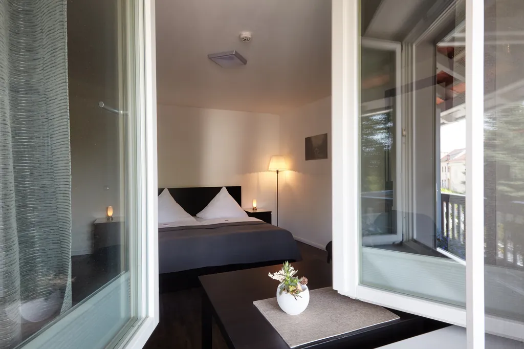 Blick durch das offene Fenster auf das Bett mit Kissen und Blumenvase