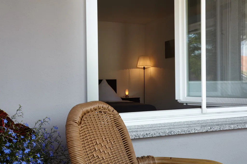 Korbstuhl vor dem Fenster, dahinter das Bett mit Nachttischlampe